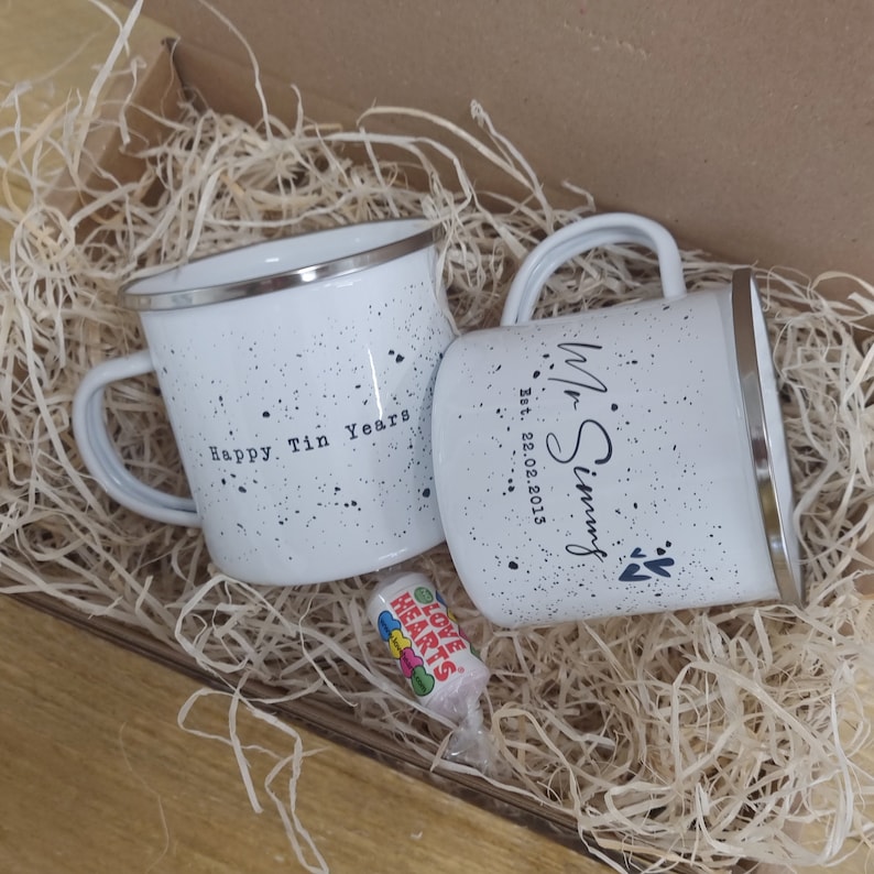 May include: Two white enamel mugs with black speckles and silver rims, nestled in a box with straw. One mug reads "Happy Tin Years," the other "Mr. Simms Est. 22.02.2013." A small candy roll with "Love Hearts" text is also in the box.