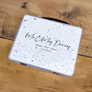 May include: A white, rectangular tin with a black handle and silver trim. The tin is decorated with black speckles and features the text "Mr & Mrs Davies" and "Happy "Tin" Years 10.16.2014."