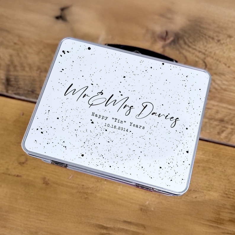 May include: A white, rectangular tin with a black handle and silver trim. The tin is decorated with black speckles and features the text "Mr & Mrs Davies" and "Happy "Tin" Years 10.16.2014."