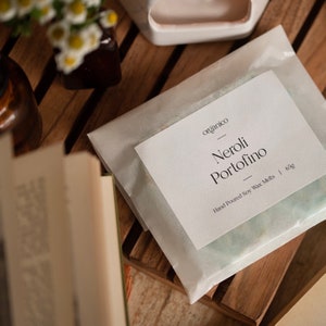 May include: A white paper bag with a label that reads "organico" and "Neroli Portofino" in black text. The label also says "Hand Poured Soy Wax Melts | 63g".