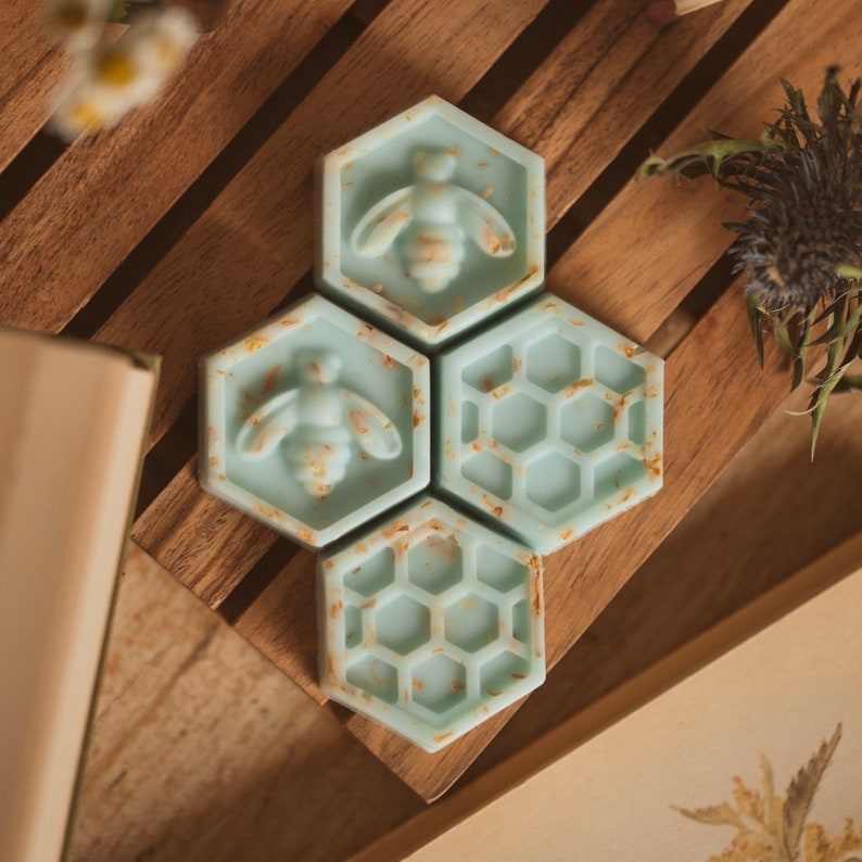 May include: Four hexagonal shaped teal blue wax melts with a honeycomb pattern and a bee design. The wax melts are arranged in a cluster on a wooden surface.