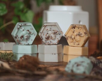 May include: A collection of hexagon-shaped wax melts in various pastel colors, each featuring a bee design. The melts are stacked on top of each other, with a white ceramic diffuser in the background. The wax melts are infused with dried botanicals.