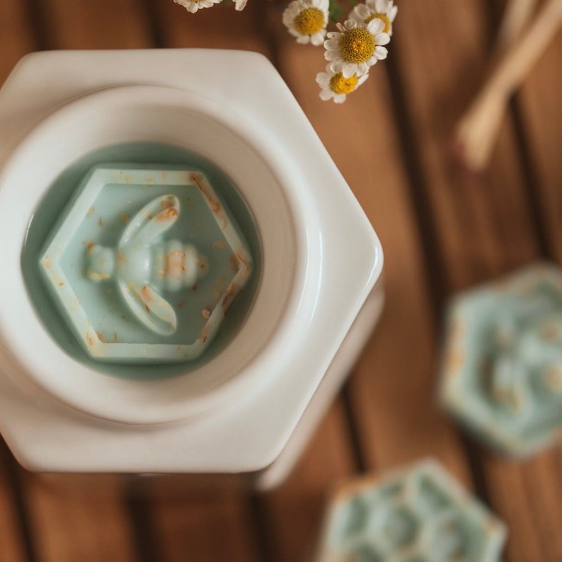 May include: White ceramic wax warmer with a hexagonal shape. The warmer has a blue wax melt with a bee design in the center. There are two additional blue wax melts with bee designs in the background.