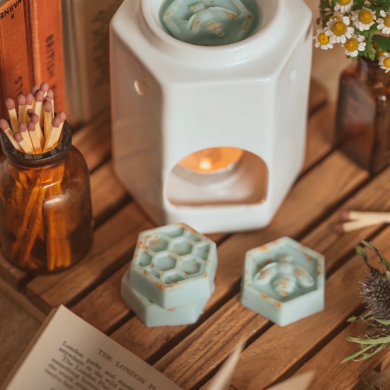 May include: Three wax melts in a honeycomb design, one with a bee, on a wooden surface with a white ceramic wax warmer and a glass bottle of matches.