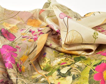 May include: A silk scarf with a botanical print featuring pink, yellow, and orange flowers, along with green foliage and beige and tan backgrounds. The scarf is folded and draped, showcasing the intricate floral design.