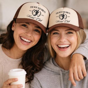 May include: Two people wearing trucker hats with a brown brim and a tan front panel. The hats feature the text "THE BOY AQUARIUM Social Club ESTD 2025" in black. One person holds a white coffee cup.