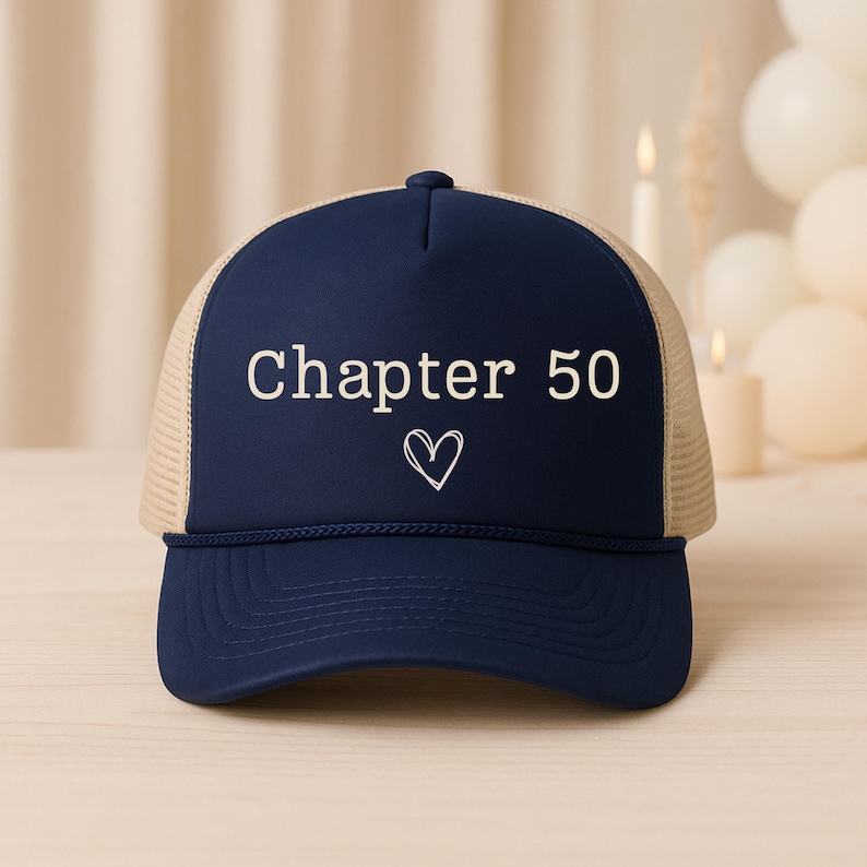 May include: A navy blue trucker hat with a beige mesh back. The front of the hat features the text "Chapter 50" in white, with a white heart symbol below. The hat is designed for casual wear.