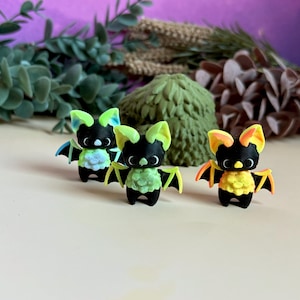 May include: Three small, black bat figurines with colourful accents. Each bat has large ears and wings with neon green, blue, and orange highlights. The bats have a textured chest and are set against a blurred background of greenery.