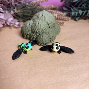 May include: Two miniature bee figurines with black wings and detailed bodies. One bee is light blue and yellow, the other is cream and black. A textured, green, tree-like sculpture is in the background.
