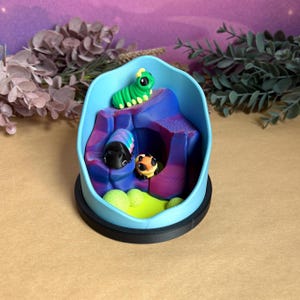 May include: A miniature diorama in a light blue oval container. It features colourful, stylised caterpillars on a purple and blue landscape. The base is black, and the background is a soft purple. The scene is reminiscent of a fantasy world.