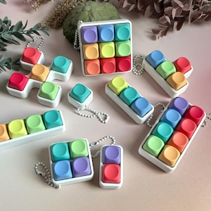 May include: A selection of rainbow-coloured fidget toys in different shapes. The toys have square, rounded buttons in shades of red, orange, yellow, green, blue, and purple. Each toy is set in a white frame and some have a chain.
