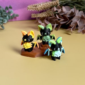 May include: Three small, colourful bat figurines. Each bat has a black body, white belly, and large ears and wings in shades of yellow, green, and blue. They are arranged on a brown, hexagonal base.