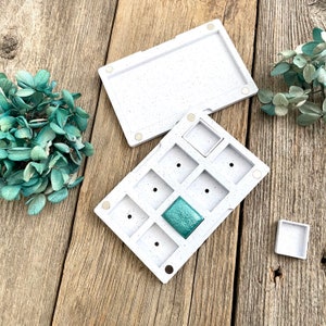 May include: A white speckled watercolour palette with a lid, featuring multiple square wells. One well contains a teal-coloured paint. A small, separate square palette is also present. The palette is open on a wooden surface.