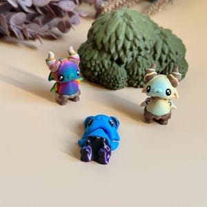 May include: Three fantasy figurines in a whimsical scene. One is rainbow-coloured with horns, another is blue with a dark blue bottom, and the third is light tan with brown accents. A green, textured, tree-like sculpture is in the background.