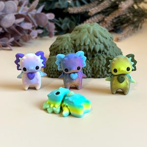 May include: A selection of vibrant, 3D-printed koala figurines. The koalas have heart-shaped designs on their chests and are in shades of blue, purple, green, and yellow. A green, textured tree-like structure is in the background.