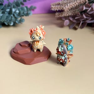 May include: Two miniature fantasy figurines. One is beige with orange floral details, the other brown with blue-green floral accents. Both have large black eyes and small wings. They are displayed on a brown, geometric base, suggesting a collectible or fantasy theme.