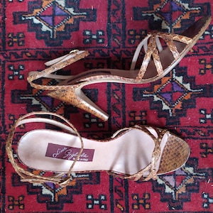 Op de afbeelding: Een paar bruine slangenleren sandalen met enkelbandjes. De sandalen hebben een kruislings bandjesontwerp en de merknaam "Lina Gigliola Made in Italy" staat gedrukt op de binnenzool.