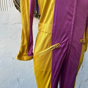 May include: A vintage jumpsuit in a color-block design. The jumpsuit features a mustard yellow and purple color scheme, with a full-length zipper and zippered pockets. The fabric has a slight sheen.