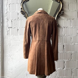 May include: A brown suede coat with a flared skirt, displayed on a marble pedestal. The coat has long sleeves and a classic collar. The background includes a decorative mirror and a textured white wall.