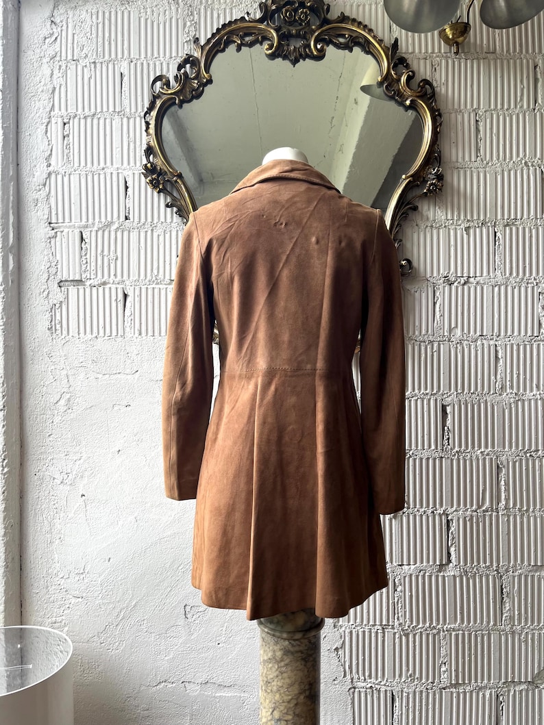 May include: A brown suede coat with a flared skirt, displayed on a marble pedestal. The coat has long sleeves and a classic collar. The background includes a decorative mirror and a textured white wall.