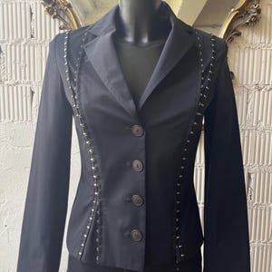 Pu&ograve; includere: Un blazer nero con collo a rever e chiusura a tre bottoni. La giacca presenta dettagli decorativi in metallo argentato lungo le cuciture. Il blazer &egrave; esposto su un manichino, mettendo in risalto la sua vestibilit&agrave; su misura e il design.