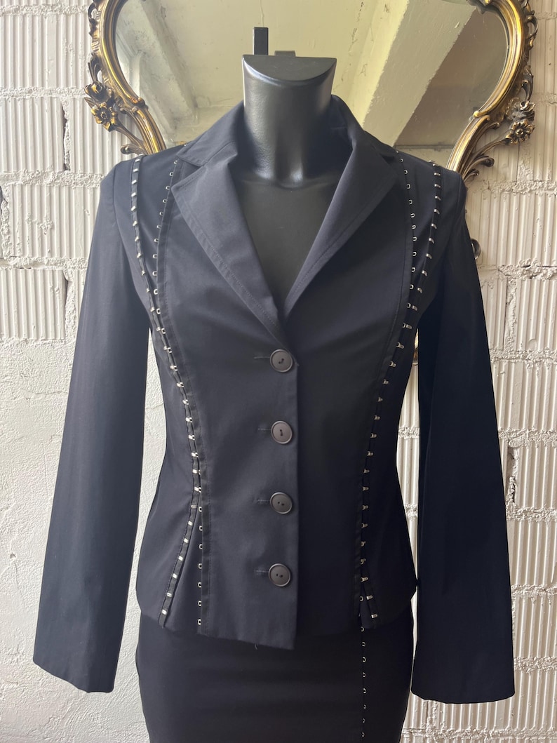 Pu&ograve; includere: Un blazer nero con collo a rever e chiusura a tre bottoni. La giacca presenta dettagli decorativi in metallo argentato lungo le cuciture. Il blazer &egrave; esposto su un manichino, mettendo in risalto la sua vestibilit&agrave; su misura e il design.