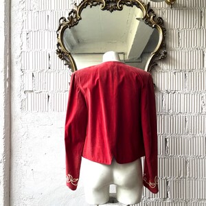 以下が含まれることがあります： 赤いベルベットのジャケット。袖口には金の刺繍が施され、マネキンに展示されています。 ジャケットは白いレンガの壁を背景に、金色の装飾鏡とヴィンテージのウォール燭台が配置されています。