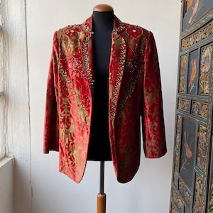 Può includere: Un blazer in velluto rosso con ricami e decorazioni dorate. Il blazer ha un collo a scialle e una chiusura a un solo bottone.