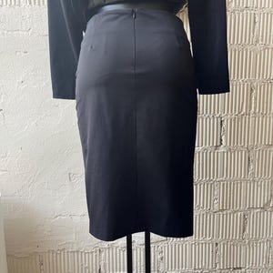 Pu&ograve; includere: Gonna a tubino nera a vita alta con cerniera posteriore. La gonna fa parte di un completo a due pezzi, con una giacca nera abbinata visibile sopra. L'outfit &egrave; esposto su un supporto in metallo nero.