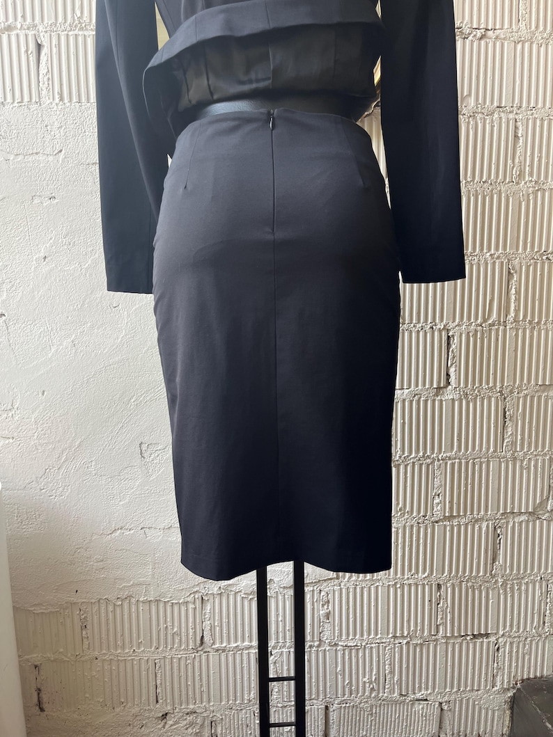 Pu&ograve; includere: Gonna a tubino nera a vita alta con cerniera posteriore. La gonna fa parte di un completo a due pezzi, con una giacca nera abbinata visibile sopra. L'outfit &egrave; esposto su un supporto in metallo nero.