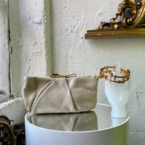 Può includere: Una pochette bianca in pelle di serpente con una chiusura e un dettaglio a fiocco dorati. La pochette è appoggiata su un tavolo bianco accanto a un braccialetto dorato su un supporto bianco a forma di mano.