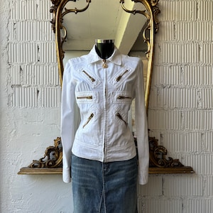 Può includere: Giacca di jeans bianca con dettagli di cerniera dorati e chiusura con bottone dorato. La giacca ha una silhouette aderente e maniche lunghe.