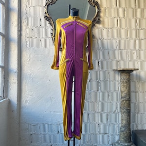 May include: A two-tone jumpsuit in gold and purple, displayed on a mannequin. The jumpsuit features long sleeves, a high collar, and a front zipper. Gold accents are present on the shoulders and pockets. The garment is made of a shiny material.