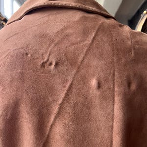 May include: Close-up of a brown suede jacket collar and upper back. The jacket has a classic collar and a soft, textured appearance. The color is a warm, earthy tone, and the fabric appears to be in good condition. The jacket is displayed on a mannequin.