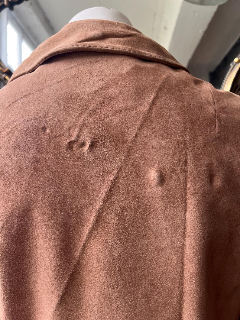 May include: Close-up of a brown suede jacket collar and upper back. The jacket has a classic collar and a soft, textured appearance. The color is a warm, earthy tone, and the fabric appears to be in good condition. The jacket is displayed on a mannequin.