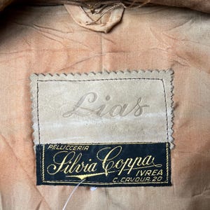 May include: Close-up of a tan garment interior with a label. The label has the name "Lias" embossed on a light tan patch. Below, a black label with gold lettering reads "Silvia Coppa" and "Pellicceria Ivrea C. Cavour 20".