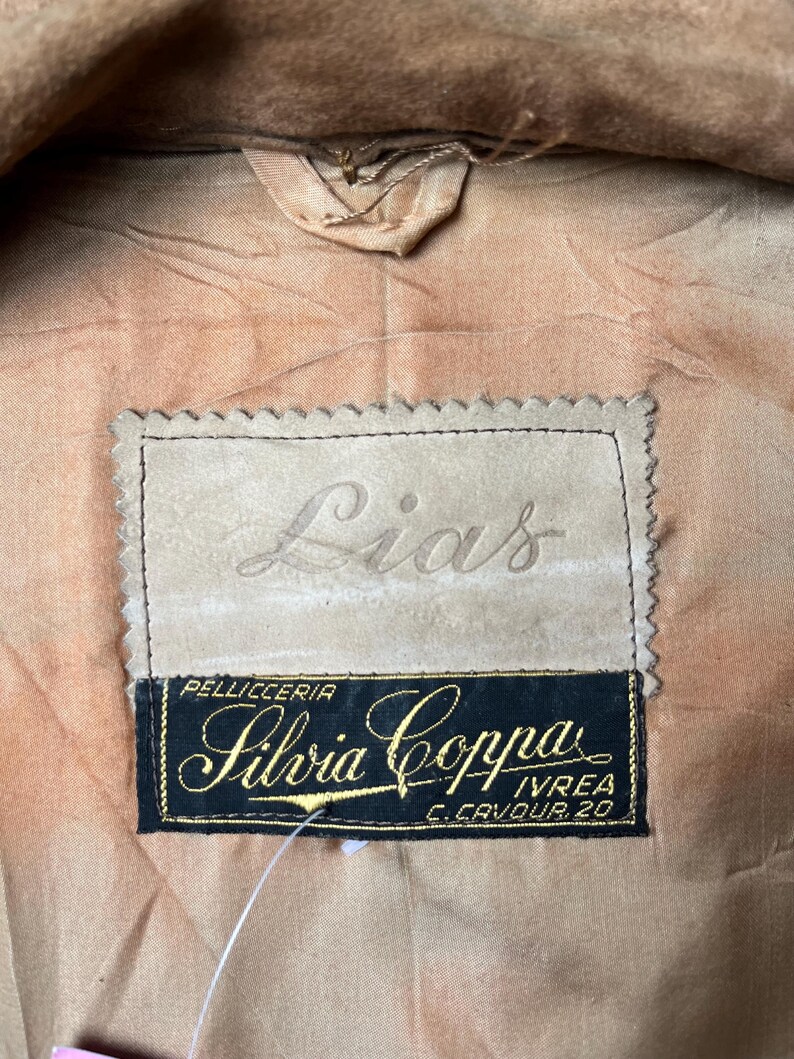May include: Close-up of a tan garment interior with a label. The label has the name "Lias" embossed on a light tan patch. Below, a black label with gold lettering reads "Silvia Coppa" and "Pellicceria Ivrea C. Cavour 20".