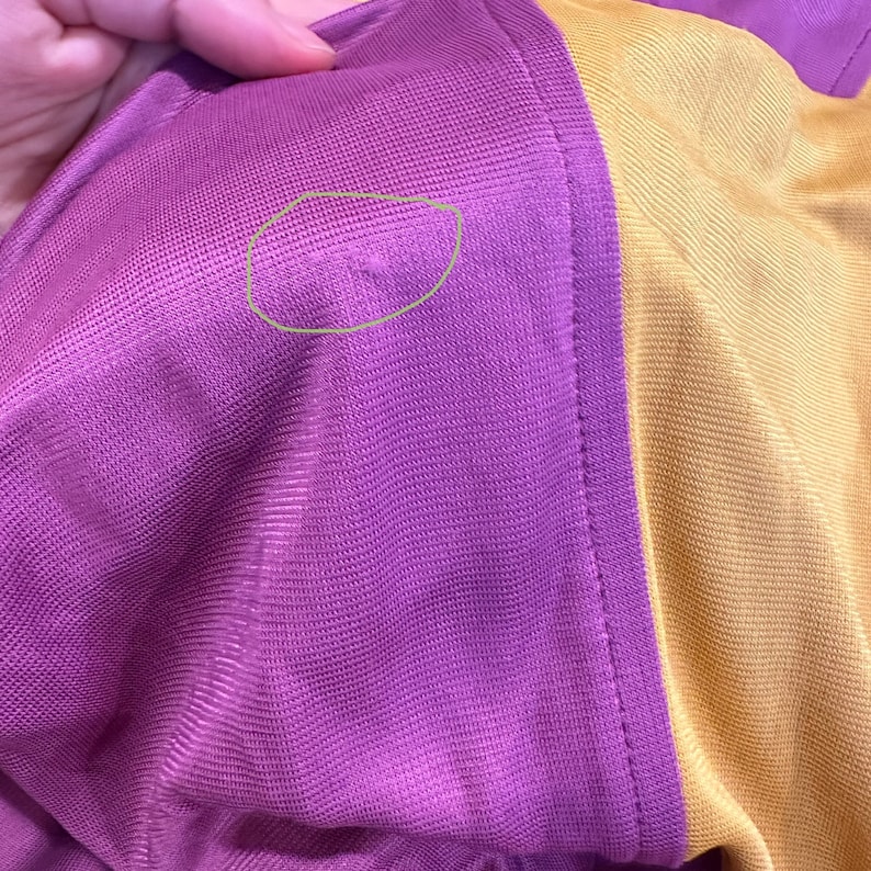 May include: Close-up of a section of fabric, showcasing a color-blocked design. The fabric is divided into three vertical sections: a deep purple, a golden yellow, and a purple border. The texture appears to be a fine mesh or knit, with visible stitching.