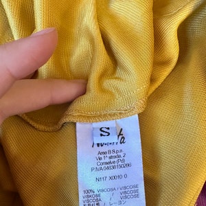 May include: Close-up of a mustard yellow garment with a small white tag. The tag has the letter "S" and text in multiple languages. The garment is made of a lightweight, possibly knitted fabric.