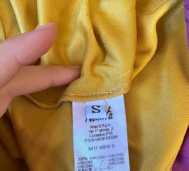 May include: Close-up of a mustard yellow garment with a small white tag. The tag has the letter "S" and text in multiple languages. The garment is made of a lightweight, possibly knitted fabric.
