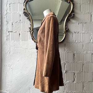 May include: A brown suede coat displayed on a mannequin. The coat has a button closure and a flared skirt. The mannequin is positioned in front of a gold-framed mirror. The background is a white brick wall.