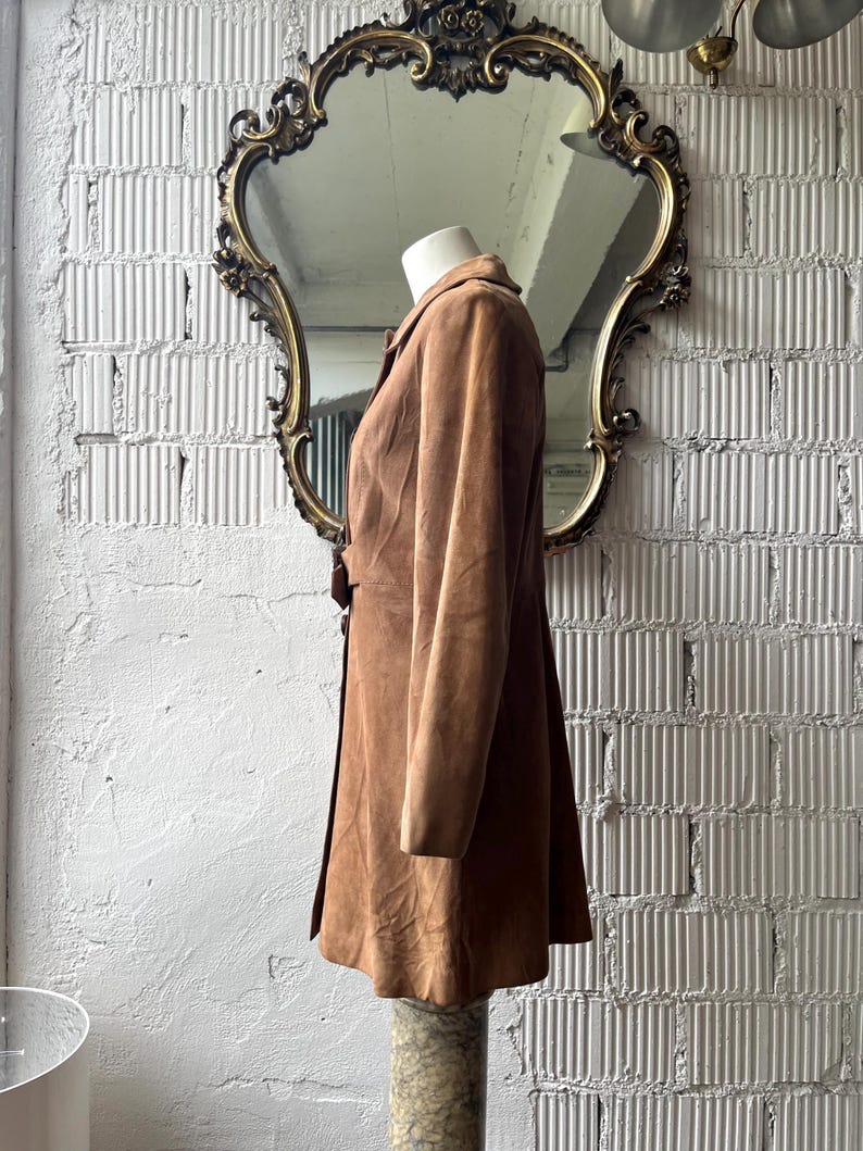 May include: A brown suede coat displayed on a mannequin. The coat has a button closure and a flared skirt. The mannequin is positioned in front of a gold-framed mirror. The background is a white brick wall.