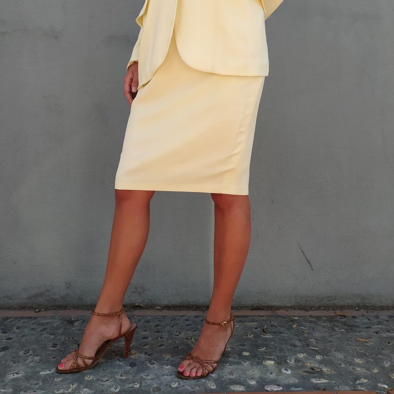 Puede incluir: Una mujer con un traje de falda amarillo p&aacute;lido y sandalias de tac&oacute;n marr&oacute;n. La falda es una falda l&aacute;piz y la chaqueta es una blazer.