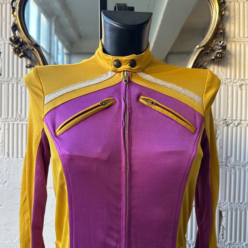 May include: Mustard yellow and purple zip-up jacket with a stand-up collar. The jacket features a silver stripe across the chest and two zippered pockets. The jacket is displayed on a black mannequin head.