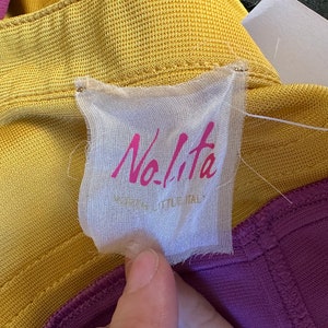 May include: A close-up of a clothing tag with the brand name "Nolita" in pink script. The tag is attached to a yellow and purple garment. The text "MADE IN LITTLE ITALY" is below the brand name.