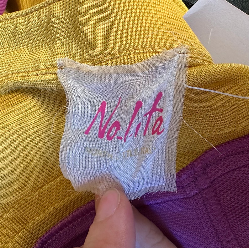 May include: A close-up of a clothing tag with the brand name "Nolita" in pink script. The tag is attached to a yellow and purple garment. The text "MADE IN LITTLE ITALY" is below the brand name.