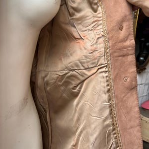 May include: A light brown suede jacket with a satin lining. The jacket has a decorative trim along the edges and button closures. The interior lining is a light tan color.