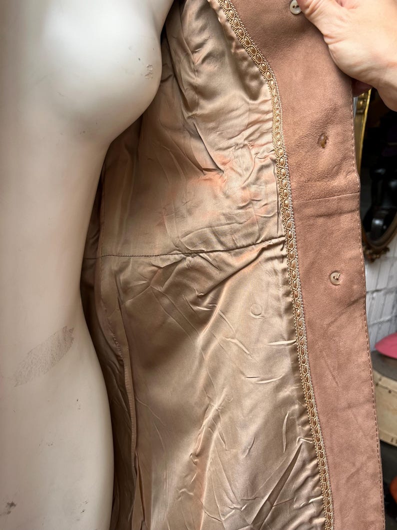 May include: A light brown suede jacket with a satin lining. The jacket has a decorative trim along the edges and button closures. The interior lining is a light tan color.