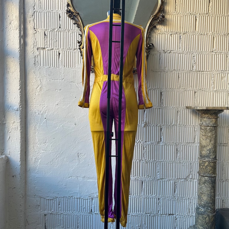 May include: A gold and purple jumpsuit with long sleeves and a high collar. The jumpsuit has a vertical purple panel down the center of the front and back. White trim accents the sleeves and collar. The garment is displayed on a mannequin.