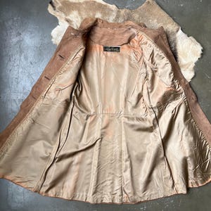 May include: Open view of a vintage brown suede jacket with a tan satin lining. The jacket has a classic collar, button closures, and a tailored fit. The lining has a label with text. The jacket is laid out on a textured surface.